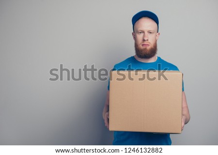 Portrait of nice handsome attractive minded bearded deliver in blue uniform holding in hands big carton box isolated over gray pastel background