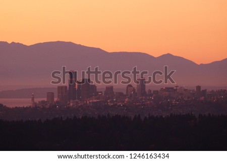 Beautiful orange sky & purple mountains highlight spectacular sunset view of downtown Seattle from city of Newcastle to the east. City buildings center frame with evergreens in silhouette at bottom.