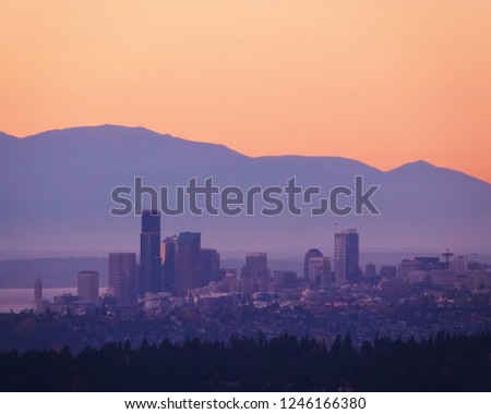 Colorful Seattle sunset. Downtown buildings in middle frame with purple Olympic Mountains rising high in background under a clear bright orange sky. Lower frame filled with evergreens in silhouette.