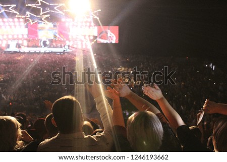 Photo colorful concert.People raise their hands and wave to the music.There are a lot of people at the stadium.On stage, colored lighting.
