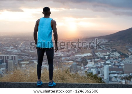 Horizontal view of attractive male dressed in casual clothes, has active run near mountains, stands back, looks attentively at beautiful sunrise at dawn, breathes fresh air, feels freedom, free space