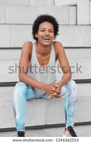 Photo of cheerful dark skinned youngster with crisp black hair, smiles broadly, dressed in casual vest and leggings, sits at steps, enjoys healthy lifestyle, favourite sport game. Vitality, wellness
