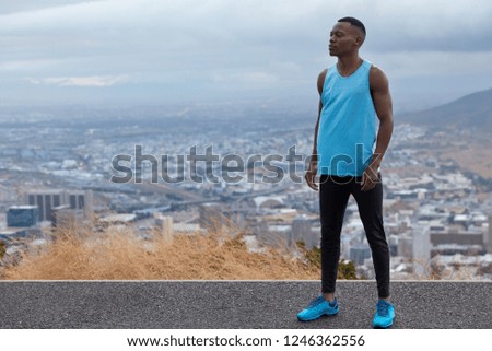 Photo of black sportsman wears blues sneakers, vest and leggings, models against altitude above horizon, big city and mountains in background, free space for your information. Panoramic view