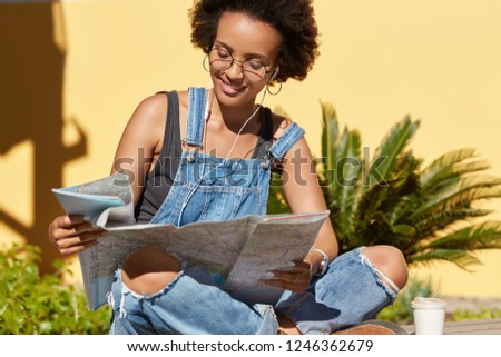 Photo of black lady with crisp hair uses destination map, searches interesting places to visit, likes sightseeing in unknown city, poses in lotus pose against tropical plants, enjoys listening music