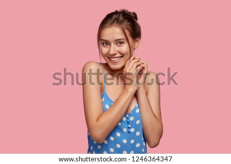 Positive content woman with joyful expression, keeps hands together, looks happily, expresses good emotions, hears something pleasant, dressed in casual outfit, isolated over pink background