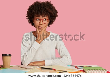 Doubtful student studies art, spends time on making sketches, holds chin and purses lips with hesitation, ponders on what to do, looks with indecisive expression aside on copy space, isolated on pink Royalty-Free Stock Photo #1246364476