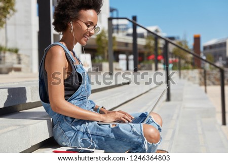 Sideways shot of black satisfied young woman checks email box on laptop computer, reads news, wears ragged denim dungarees, sits on steps against city background, relaxes outside in summer day