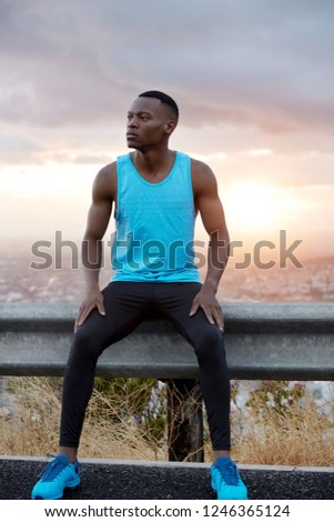 Photo of thoughtful black man in blue vest, black trousers and tariners, poses at road sign over wonderful sunrise view, has jogging exercise at morning before work. Vertical shot. Endurance