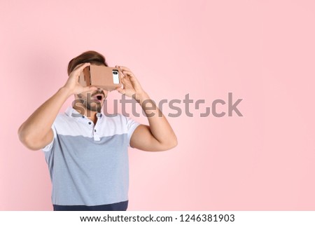 Young man using cardboard virtual reality headset on color background. Space for text