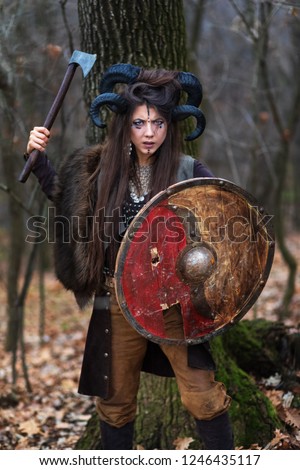 Outdoor portrait of young beautiful viking woman warrior with blue eyes wearing ram horns holding ax and shield looking threatening. Female Viking on forest with specific makeup and fur collar