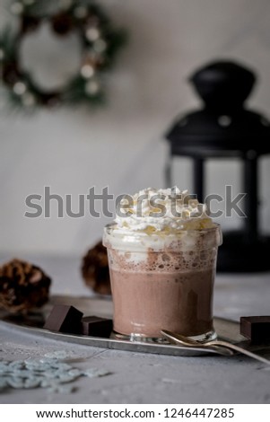 Sweet tasty hot chocolate with cream in glass on metal tray with spoon, black lantern,new year wreath, cones and slice of dark chocolate near at gray concrete background. Christmas composition.