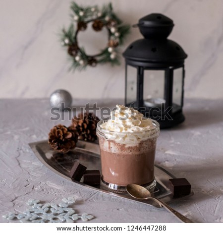 Sweet tasty hot chocolate with cream in glass on metal tray with spoon, black lantern,new year wreath and cones near at gray concrete background. Christmas composition.
