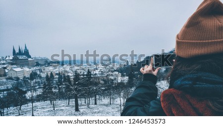 A young girl taking a picture of the snow covered city of Prague from the mountains.