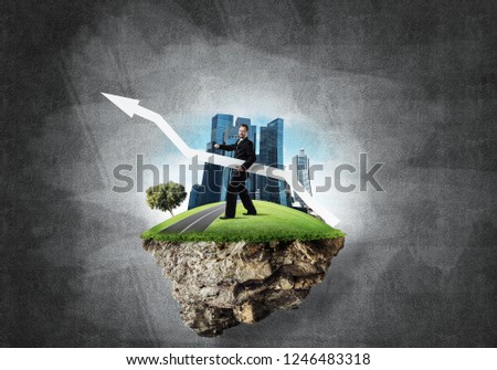 Conceptual image of young and confident business man in suit holding big white arrow in hands while standing on flying island against gray concrete background