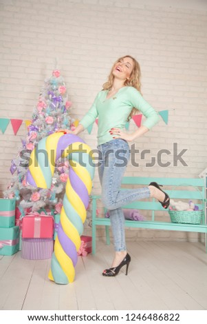 Pretty Girl Wearing Blue Sweater and Jeans Having Fun In Her White Room Decorated For Christmas Holidays With Huge Candy Sweets and Cute Xmas Tree With Bows