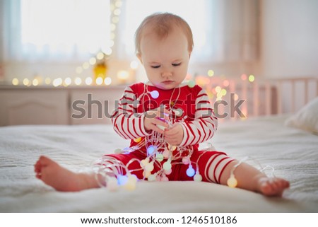 Happy little baby girl wearing pyjamas playing with light garland on her very first Christmas. Celebrating Xmas with kids at home
