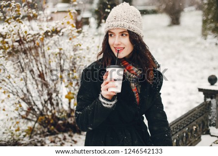 young and stylish girl in black coat and white hat drinking coffee in winter city