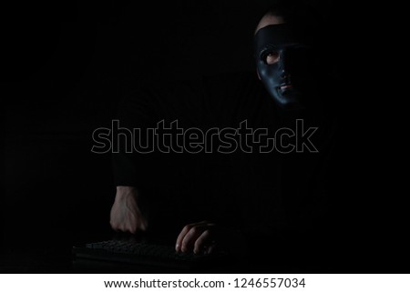 A man in a mask prints on the keyboard at a table in the dark
