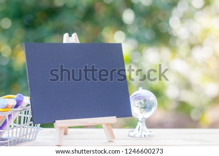 Image of empty small chalkboard on wooden easel, colorful chalk sticks in basket and globe over wooden table outdoor with blurred nature green background. Mock-up of menu blackboard