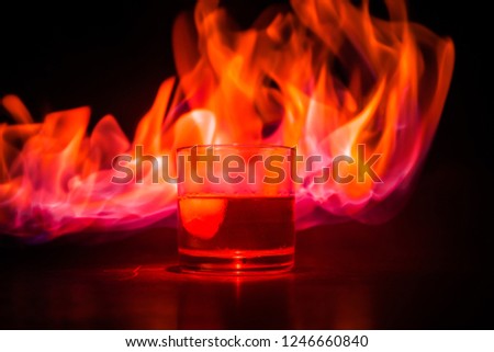 Whiskey in fire concept. Glass of whiskey and ice on wooden surface with color light and fog on background. Close up. Selective focus