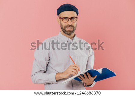Pleased attractive man with stubble, wears transparent glasses, fashionable hat and shirt, writes with pen in notepad, prepares for speaking in front of auditorium, isolated over pink background Royalty-Free Stock Photo #1246686370