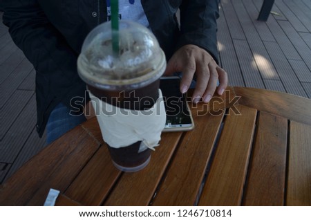 A glass of iced coffee Americano wrapped with offwhite tissue paper inserted with green straw, placed on a wooden slat table while a man sitting and sliding his finger on a touchscreen mobile phone