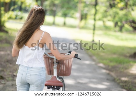 Rear shot of Asian woman wheel or push bicycle by hands to bike lanes at park with foliage bokeh background. People Healthy and sport lifestyle concept with copy space for text.