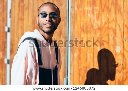 Young black man wearing casual clothes and sunglasses, smiling against a wooden background. Millennial african guy with bib pants outdoors
