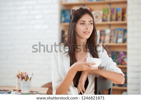 Adorable young woman posing with cup of coffee or tea in hand. Gorgeous girl looking away and sitting. Model with chestnut hair wearing in white shirt. Painter in art studio.