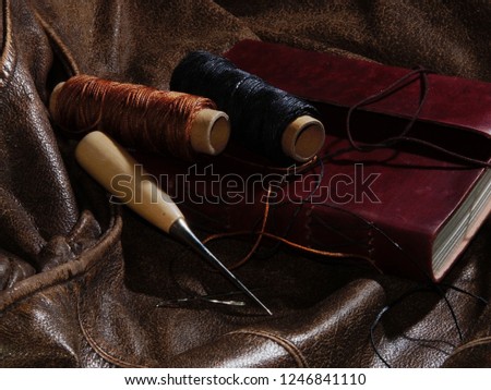 leather working supplies: waxed thread, needles and a stitching awl are placed with a hand made leather bound journal on an aged leather backdrop.