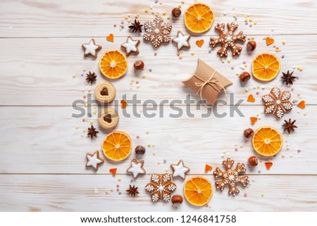 Christmas holiday  wreath  background with gift box, homemade cookies and gingerbread,  dry orange slices, nuts, spices and cones on white wooden background. Top view, close up