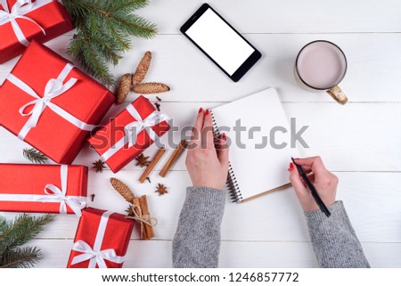 Woman writing New year goals (to do list) to notebook on desk with Christmas decorations, red gift boxes, cup of cocoa or chocolate and mobile cellphone with blank screen, copy space. Top view