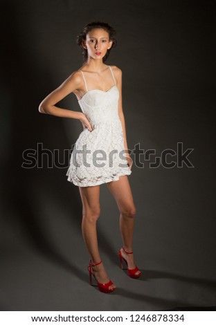 Slim Teen Brunette in a white dress with red shoes on a dark grey background with hard directional light camera right and softer backlight