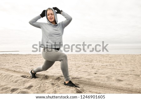 Wellness, energy, health and activity concept. Picture of athletic young blonde woman with perfect muscular body standing in lunge pose on beach, smiling broadly, enjoying training outdoors