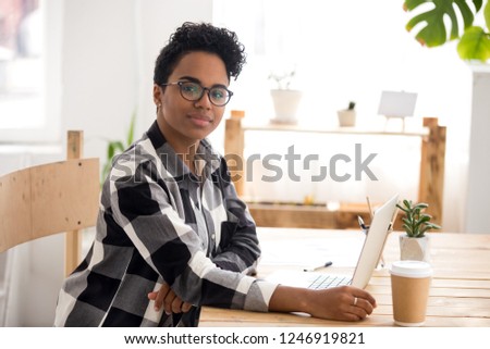 Portrait of beautiful African American female in glasses working at laptop in loft office, smiling black woman look at camera posing at cozy workplace, mixed race girl sit at desk using computer