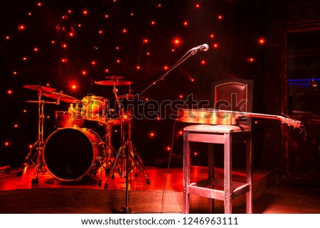 Preparation for a concert. The drum kit stands on the stage. On a high chair the acoustic guitar lies. The microphone is filled. On the stage red light shines. Dark background and fires
