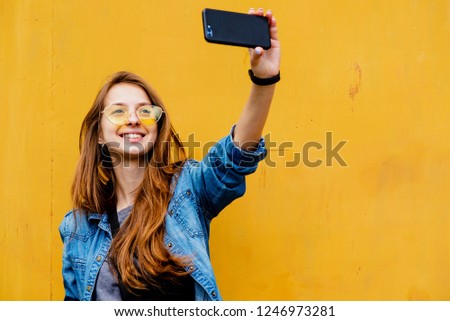 beautiful girl on a yellow background. Young girl of European appearance with long red hair in jeans clothes and yellow glasses smiles and makes a selfie on a yellow background. space. close-up