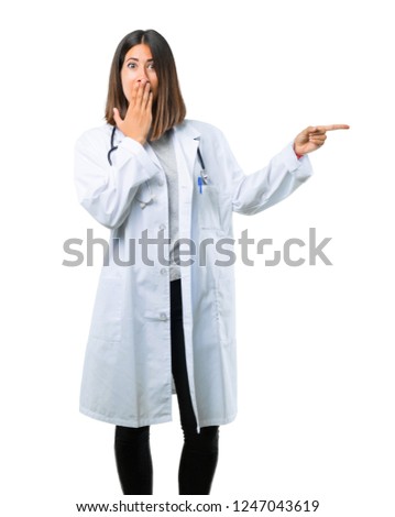 Doctor woman with stethoscope pointing finger to the side and presenting a product with a surprised face while covering mouth on isolated white background