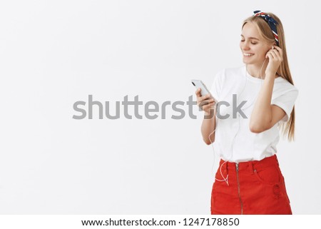 Studio shot of joyful modern young female in red skit and headband standing half-turned while putting on earphone and smiling satisfied at smartphone screen getting ready to go for walk with music