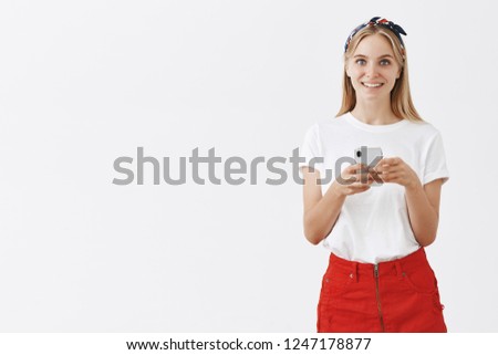 Indoor shot of happy good-looking blonde female in red skirt and headband looking happily at camera holding smartphone making notes during interview with gadget over gray background