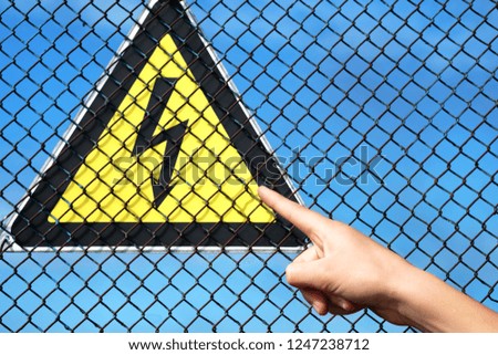 hand pointing to the sign of high voltage, before the sign of the metal grid behind the sign of the blue sky