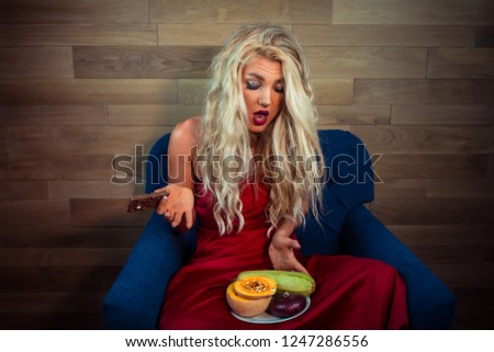 Young woman in red dress sits on armchair with chocolate in hand and looking on vegetables. Funny housewife choosing between healthy or unhealthy food