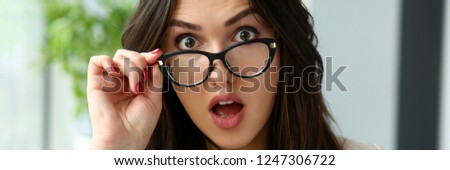 Young beautiful business woman holding glasses with her hand and opened her mouth to astonishment looking at the camera