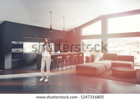 Woman in corner of gray living room with concrete floor, gray sofa standing near round coffee table and kitchen in the background. Toned image