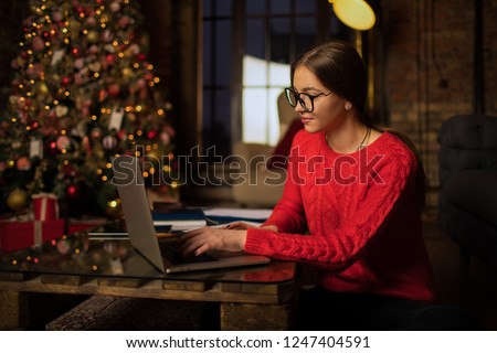 Woman freelancer searching information via netbook while sitting in loft interior near beautiful Christmas tree in winter holidays. Female having distance work via laptop computer, sitting in home 