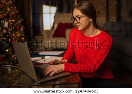 Hipster girl in stylish glasses professional fashion magazine editor checking e-mail on laptop computer while sitting in home interior near Christmas tree during holidays in winter. Online learning 