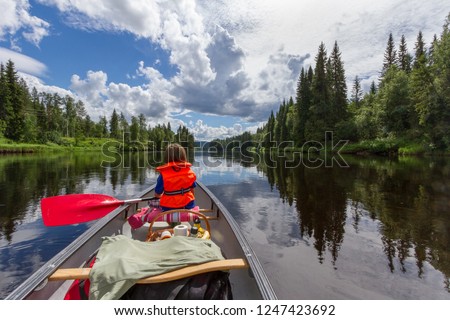A boy canoe paddling on a calm river with the surrounding forest and cloudy sky reflecting in the the water in beautiful northern Sweden  Royalty-Free Stock Photo #1247423692