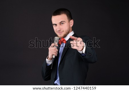 Handsome Master of ceremonies in black suit holding microphone in hand on black background. Showman, tv