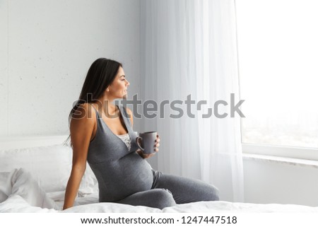 Image of an amazing healthy pregnant woman indoors at home sitting posing drinking tea or coffee on bed.