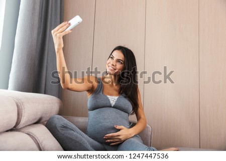 Image of an amazing healthy pregnant woman indoors at home sitting on sofa make a selfie by phone.